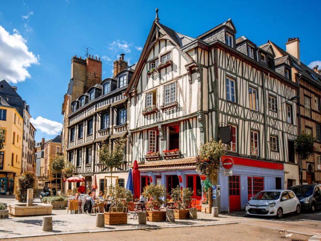 A sunny street scene in Rouen, France, featuring half-timbered houses with traditional architecture. The buildings have multiple stories with visible wooden beams and white fill. The ground floor of the closest building has a red storefront and outdoor seating arranged in front, suggesting a cafe or restaurant. There is a stone fountain in the foreground, and the clear blue sky suggests a pleasant day. Several potted plants add greenery to the scene, and a few cars are parked along the side of the street.