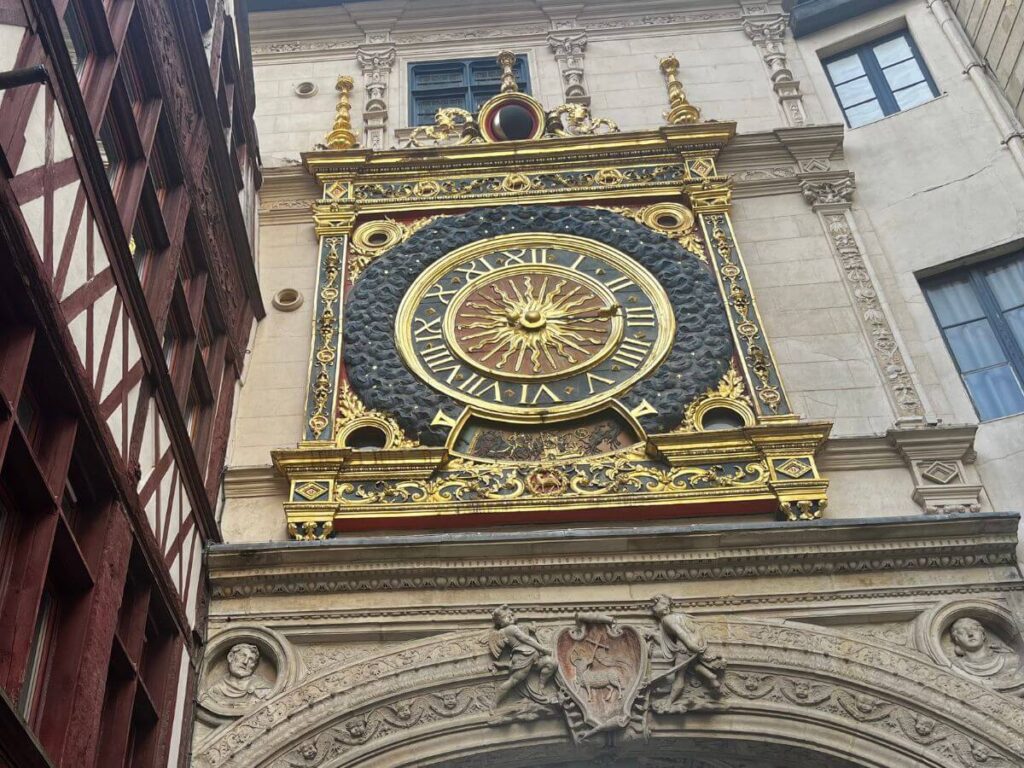 This image features the Gros Horloge, an iconic astronomical clock in Rouen, France. The clock face is ornate, with gold embellishments and Roman numerals, set against a dark, detailed background. Above the clock is a small golden sun sculpture, and the entire piece is framed by an elaborate arch with classical architectural details. To the left, the edge of a half-timbered building is visible, contrasting with the clock's grandeur.