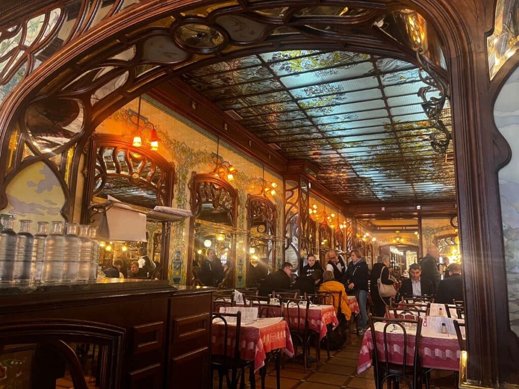 The ornate interior of Bouillon Chartier Montparnasse, showcasing an Art Nouveau design with intricate woodwork, decorative mirrors, and a stained glass ceiling. Diners are seated at tables covered with red and white checkered tablecloths.