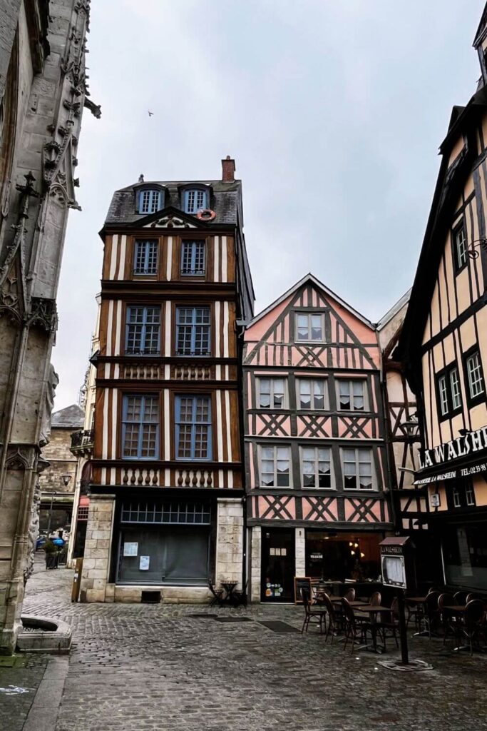 A picturesque cobblestone street in Rouen, France, flanked by distinctive half-timbered houses. The house on the left is particularly striking, with its upper stories dramatically leaning to one side, showcasing the historical settling of buildings over time. Its timber frames are filled with cream and brown colors, and it features slate grey roofing. To the right is another half-timbered building with pink and grey hues. Outdoor seating from a café spills onto the sidewalk in the foreground, inviting a moment of leisure. The gloomy sky above suggests overcast weather. The presence of these well-preserved medieval structures provides a window into the architectural heritage of Rouen, contributing to its charm as a historical destination.