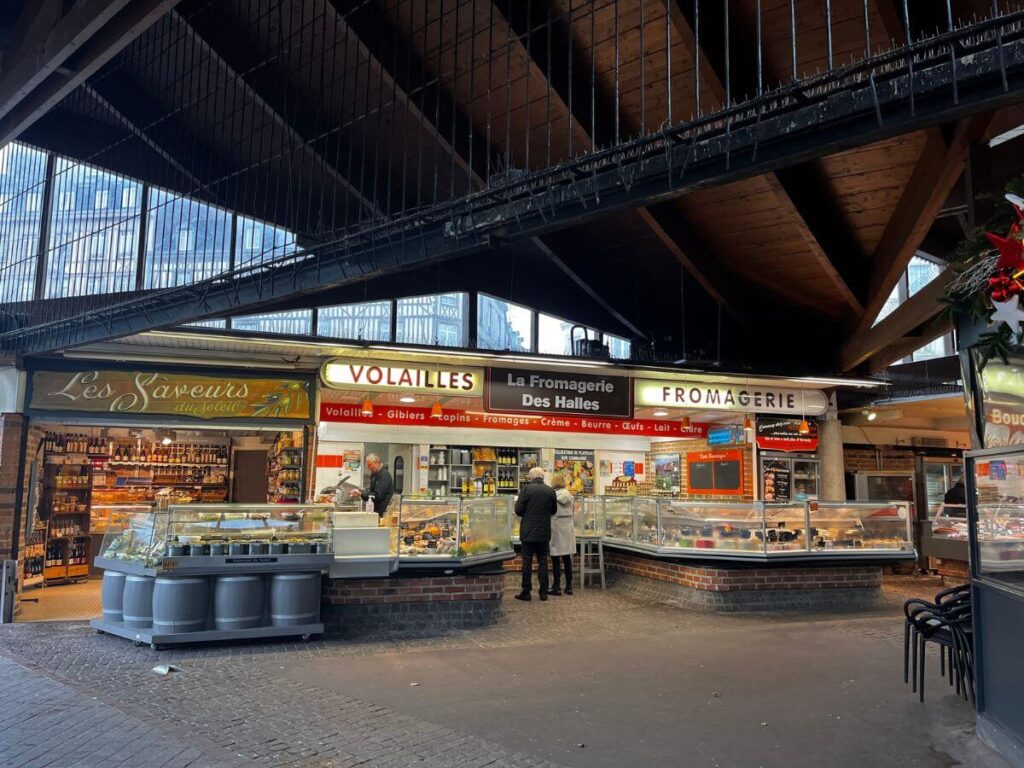 This image is taken inside the Market Place du Vieux in Rouen, showcasing a lively indoor market scene. Stalls with signs for "Volailles" and "Fromagerie" indicate a variety of poultry products and cheeses available. Patrons can be seen browsing, reflecting the daily life and culture of local shopping in Rouen. The market's interior is framed by dark wooden beams, with natural light filtering through the glass roof above.