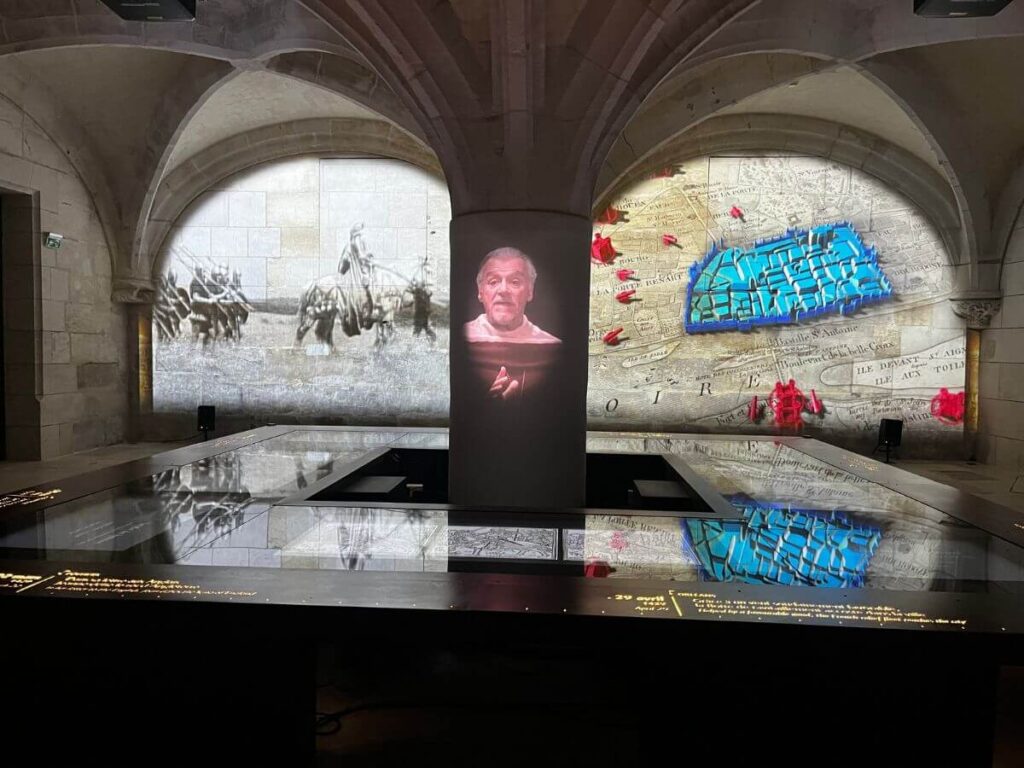 This picture shows an exhibition space within the Mythothèque in Rouen, with projections on the stone walls. Historical images and texts are illuminated, including a figure on a screen that seems to be narrating a story, bringing history to life. The room's design merges the ancient architecture with modern multimedia storytelling techniques.