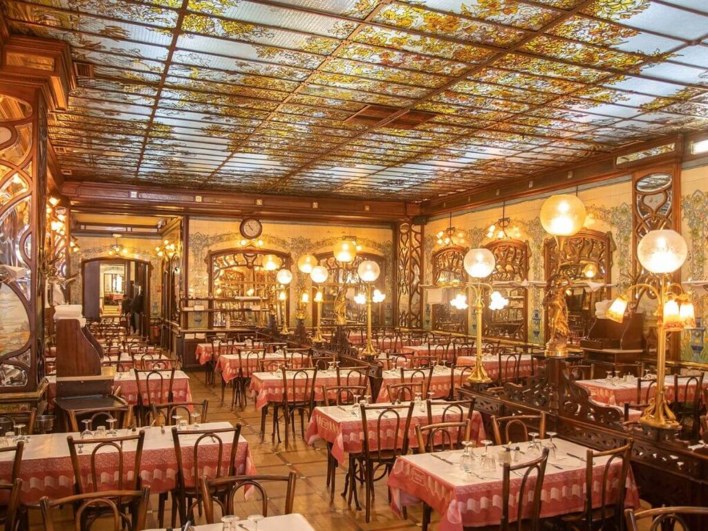 This image depicts the interior of a traditional French bouillon restaurant, showcasing an opulent Art Nouveau design. The ceiling is adorned with intricate, colorful glasswork, while the walls are decorated with ornamental mirrors and woodwork. The room is illuminated by spherical, frosted glass lamps mounted on ornate bronze stands. Red-and-white checkered tablecloths cover numerous wooden tables arranged neatly across the patterned tile floor, ready to welcome diners. The space exudes a warm, inviting atmosphere, encapsulating the charm and elegance of a bygone era in Parisian dining culture.