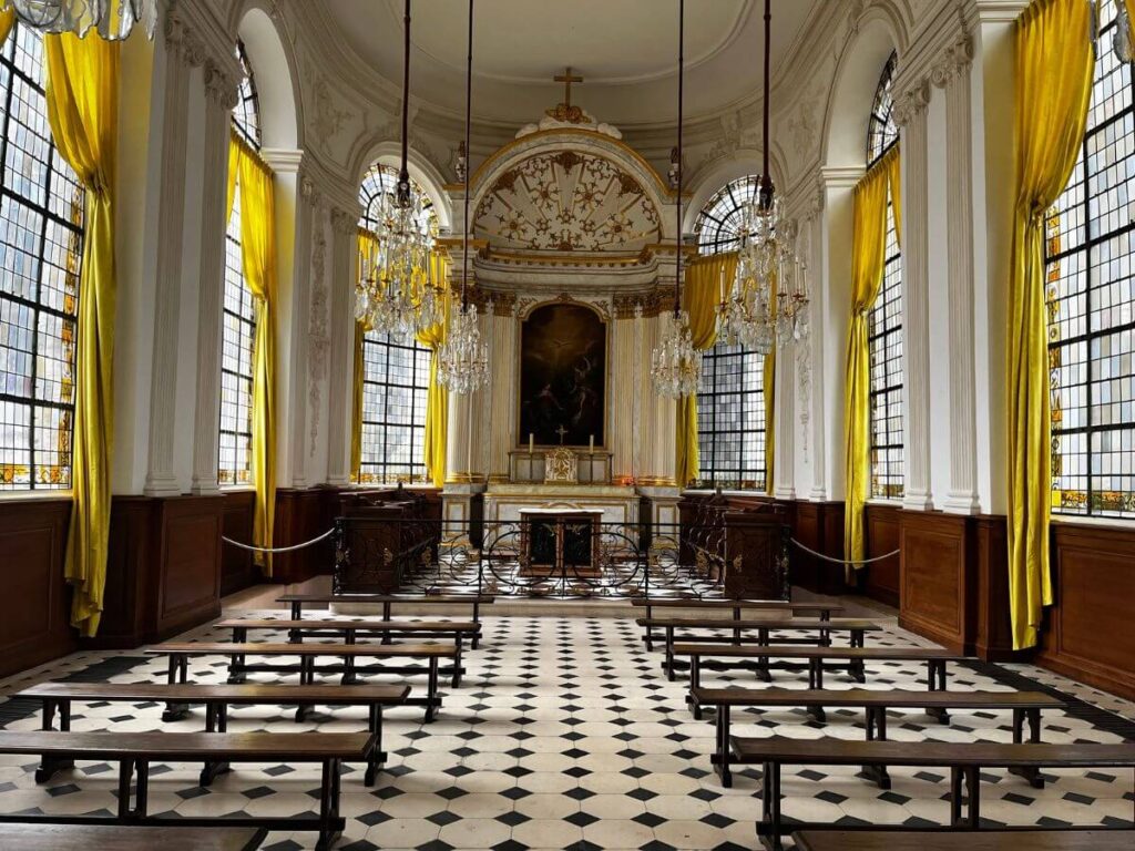 Inside the State Room in Rouen, this image reveals a grand, baroque-style interior with high ceilings, large windows with yellow drapes, and checkered black and white flooring. Crystal chandeliers hang from the ceiling, and wooden benches are neatly arranged. The room exudes elegance and historical importance, reflective of its ceremonial use.