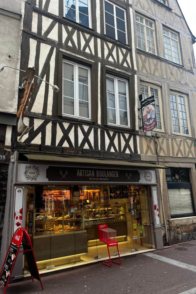 The front of a traditional French bakery, 'Artisan Boulanger,' located in a half-timbered building characteristic of some historical French towns. The upper stories of the building feature a timber frame with white infill, while the ground floor showcases a large window displaying an array of bread and pastries, illuminating the shop's interior with warm light. A red sandwich board stands on the pavement, advertising the bakery's offerings alongside a tall red chair and small table, suggesting a place for customers to enjoy a quick bite. The shop sign features a classic French baguette, adding to the authentic ambiance.