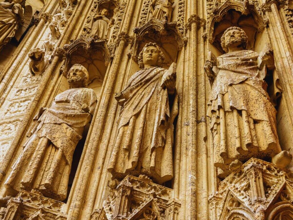 A close-up image showing the elaborate stone carvings on the exterior of Rouen Cathedral. The figures, possibly saints or biblical characters, are adorned with detailed robes and stand within ornate niches. The craftsmanship displays the weathering of time, highlighting the texture and age of the stone.
