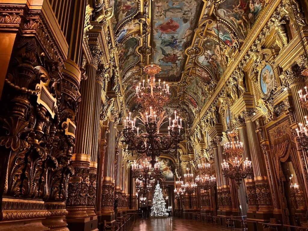 The luxurious interior of the Palais Garnier, featuring opulent golden ornaments, intricate wall carvings, and grand chandeliers, all under a beautifully painted ceiling depicting classical scenes. The perspective includes a view down the lavish hallway lined with ornate columns and wall art, with a Christmas tree visible in the distance, adding a seasonal touch to the historic venue.