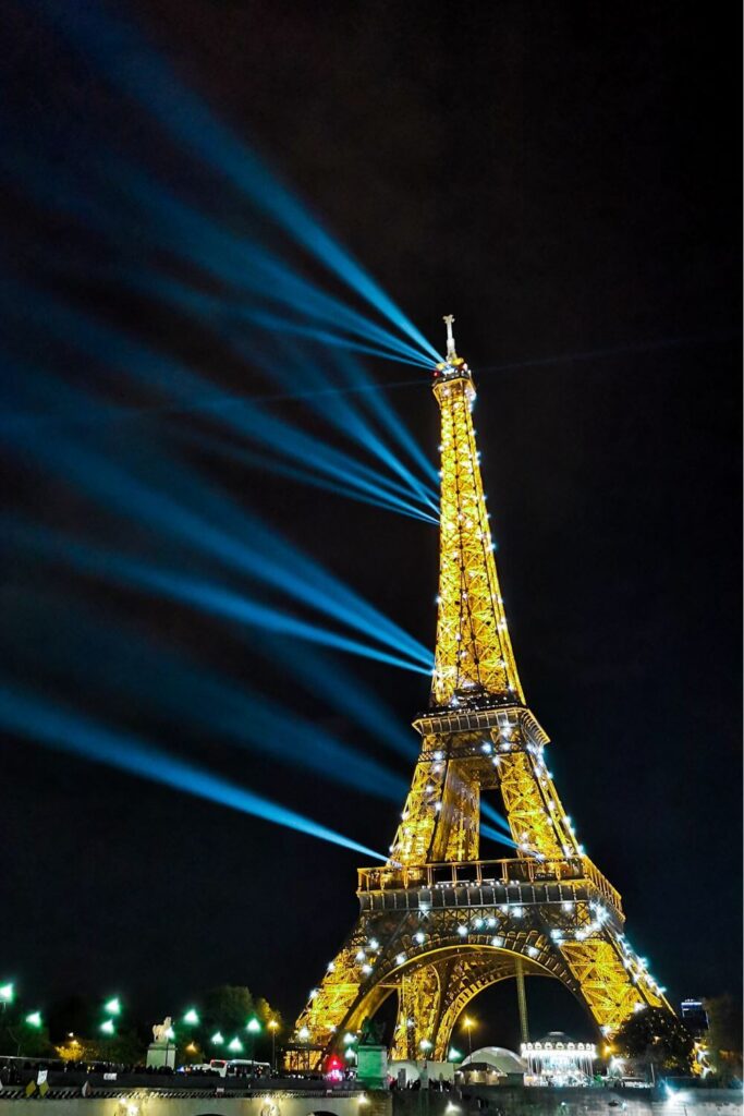 The Eiffel Tower at night, with its golden illumination casting a warm glow against the dark sky. The tower's beacon shines brightly, sending vertical beams of light into the sky, while the surrounding area is subtly lit with ambient streetlights.