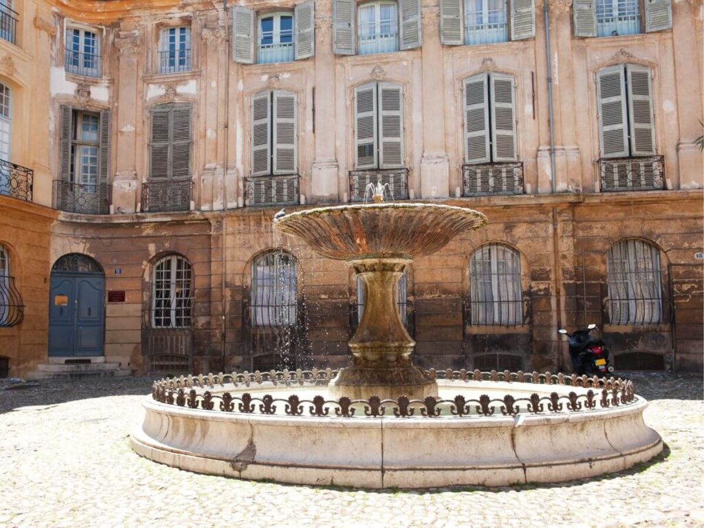 A classic stone fountain sits serenely in a cobblestone courtyard in Aix-en-Provence, a city famed for its thermal springs. The water gently cascades over the edges, surrounded by a circular railing. In the background, the weathered façade of traditional Provencal architecture, with its pale orange hues and shuttered windows, adds to the tranquil and timeless atmosphere of this historic French city.