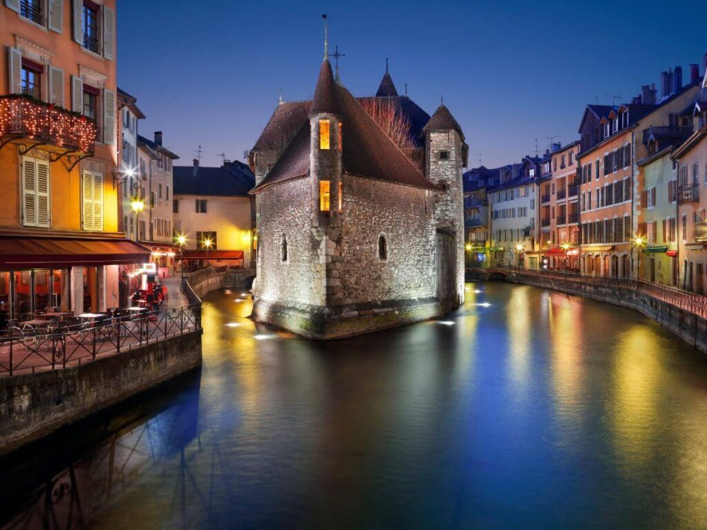 Twilight over Annecy, known as the Venice of the Alps, with its historic Palais de l'Isle jail illuminated and reflecting on the calm canal waters. Surrounding the canal, vibrant building facades with glowing windows and bustling restaurants create a cozy atmosphere, while the soft lights cast golden hues on the water, enhancing the town's picturesque charm.