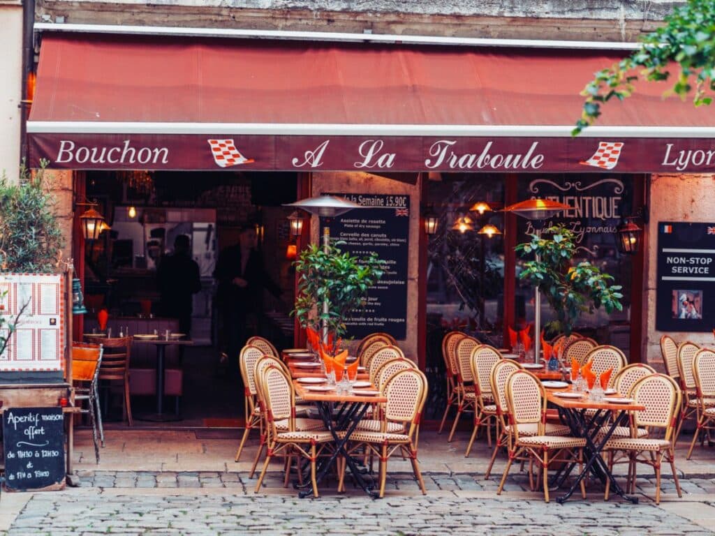GPT
A cozy Lyonnais bouchon flaunts its traditional charm with a red awning and wicker chairs invitingly arranged outside. The quaint, cobbled street and rustic façade set the scene for an authentic French dining experience.