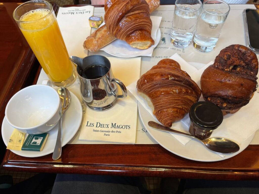 A typical French breakfast at Les Deux Magots café, featuring freshly baked croissants, a chocolate muffin, a glass of orange juice, a cup of espresso, and a pot of chocolate spread, ready to be enjoyed on a white tablecloth.