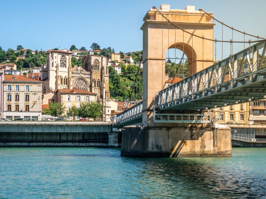 The image captures a green steel bridge foregrounding the historical architecture of the city of Vienne, with a clear focus on the ornate façade of a cathedral. The calm river beneath reflects the bridge’s structure and adds a serene quality to the urban landscape under a bright, clear sky.