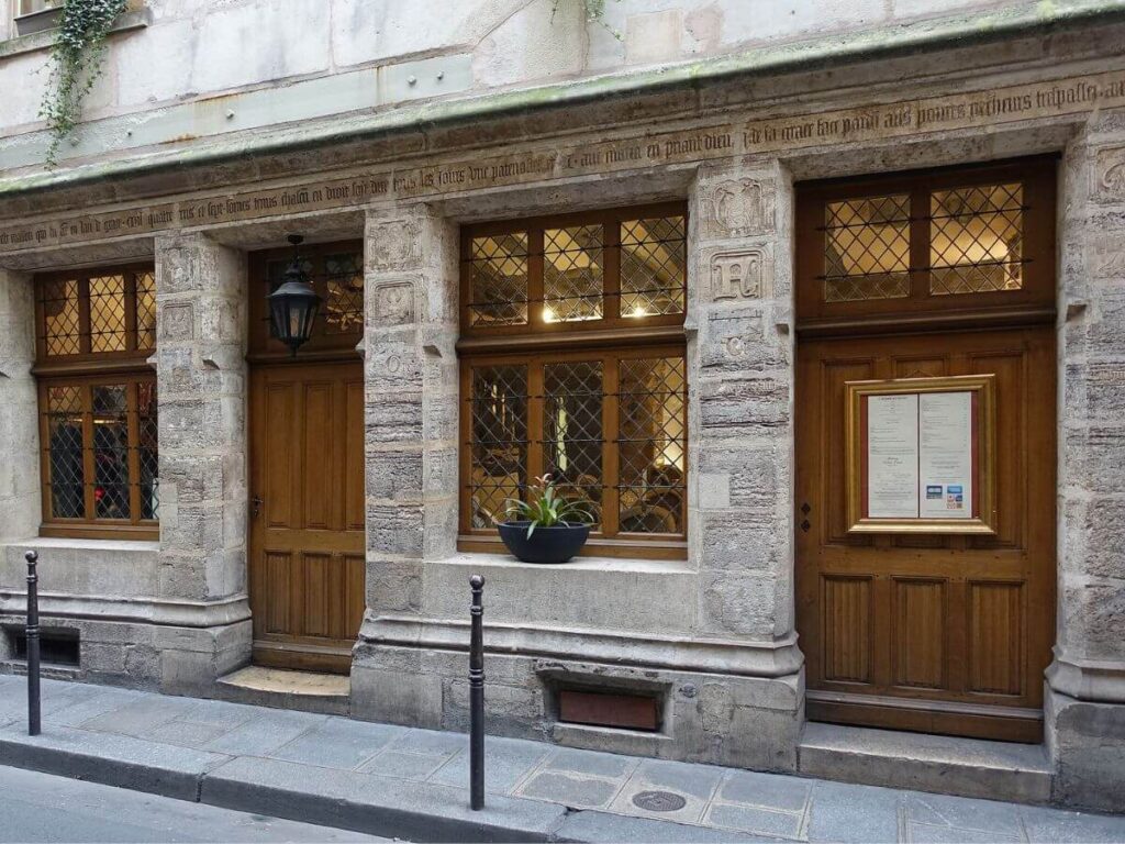 An aged stone building facade with intricate carvings and inscriptions above wooden doors and windows with diamond-patterned leaded glass. A suspended lantern and a potted plant add to the historical charm of this Parisian architecture proving that Nicolas Flamel lived in Paris