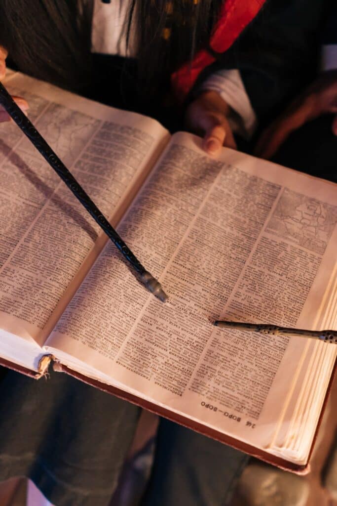 Close-up of a person reading a large, open book with a wand resting on the pages, illuminating the dense text and intricate illustrations, reminiscent of magical themes and narratives.