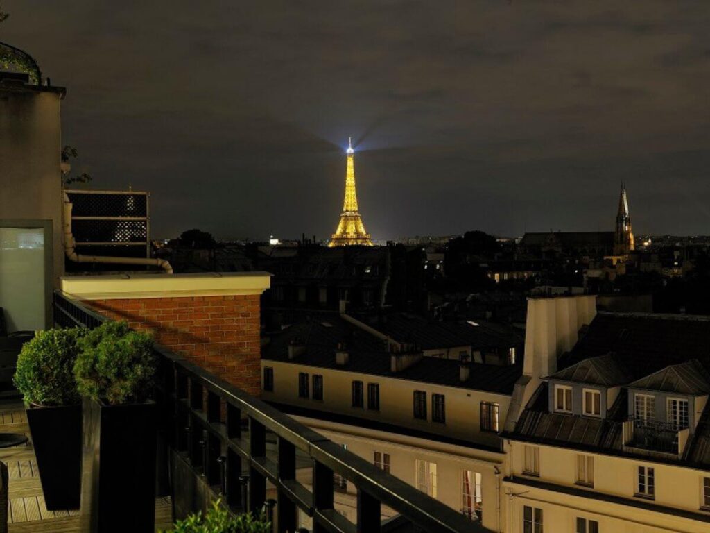 A rooftop view at night from a hotel balcony overlooking Paris, with the Eiffel Tower illuminated in the distance. The city's historic architecture is visible under a dusky sky, with scattered lights from buildings adding to the urban ambiance.