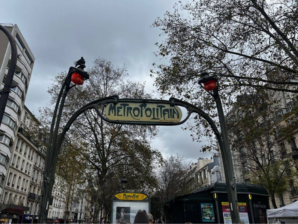 Iconic Art Nouveau entrance of the Paris Métro with the 'Metropolitain' sign and red signal lamps, against a backdrop of cloudy sky and leafy trees.