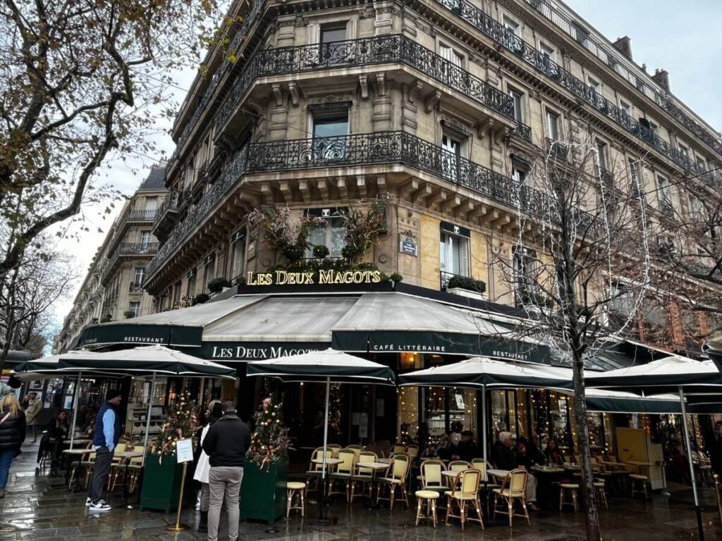The iconic Les Deux Magots café in Paris, with its traditional green awning and outdoor seating, bustling with patrons on a cloudy day, as bare trees hint at the winter season.
