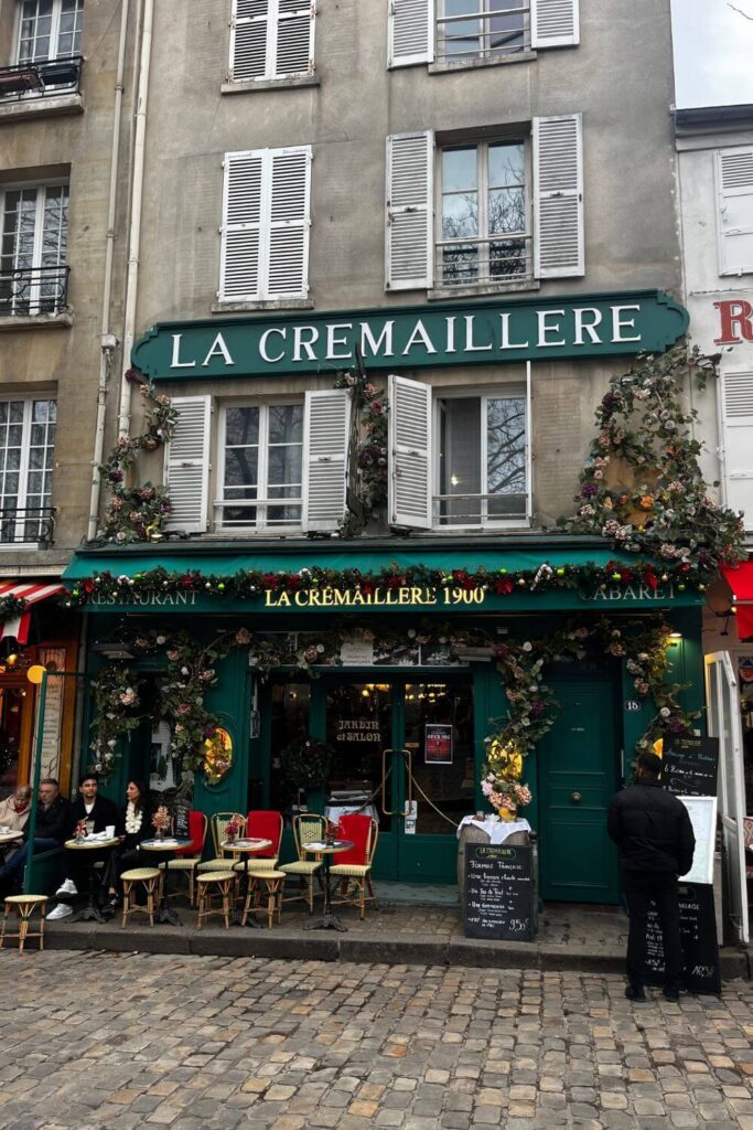 The quaint facade of 'La Cremailiere 1900' restaurant in Montmartre, Paris, with patrons dining outside and the entrance adorned with floral decorations.