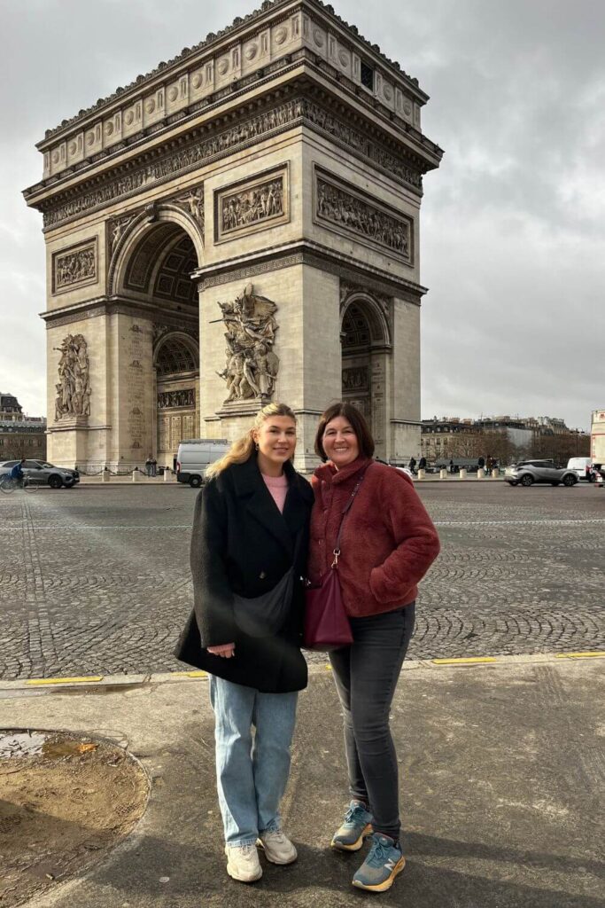 Two people smiling in front of the Arc de Triomphe in Paris, with intricate sculptures adorning the monumental arch.