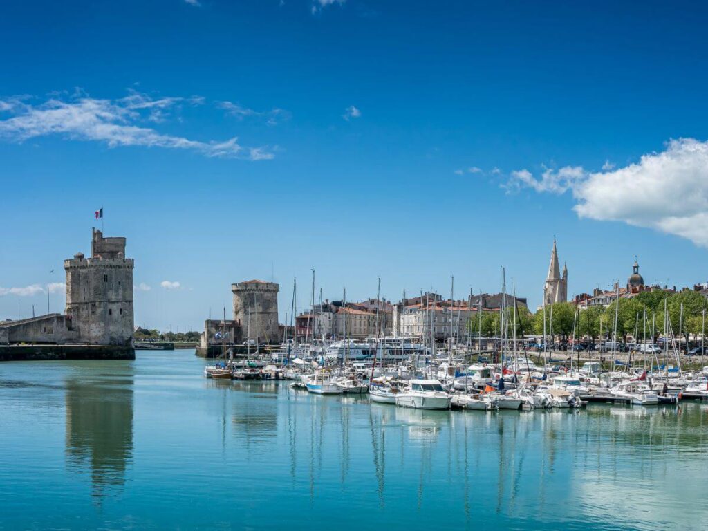 A tranquil marina in La Rochelle, France, where a multitude of sailboats are moored in calm turquoise waters. In the background stand the iconic towers guarding the entrance to the old port, under a vast blue sky dotted with wispy clouds, reflecting the city's historical significance and its maritime heritage.