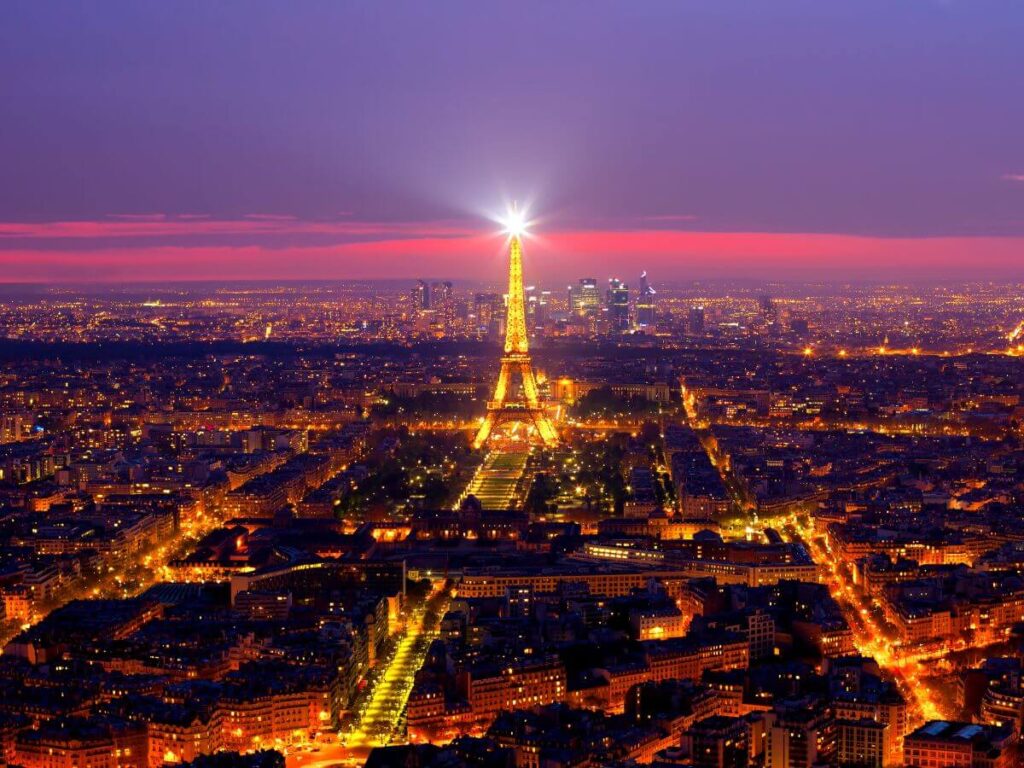 A panoramic nighttime view from the Montparnasse Tower observation deck, showcasing the Eiffel Tower radiating light amidst the Parisian cityscape. The sky transitions from twilight blues to deep sunset reds, while the urban grid below sparkles with streetlights.
