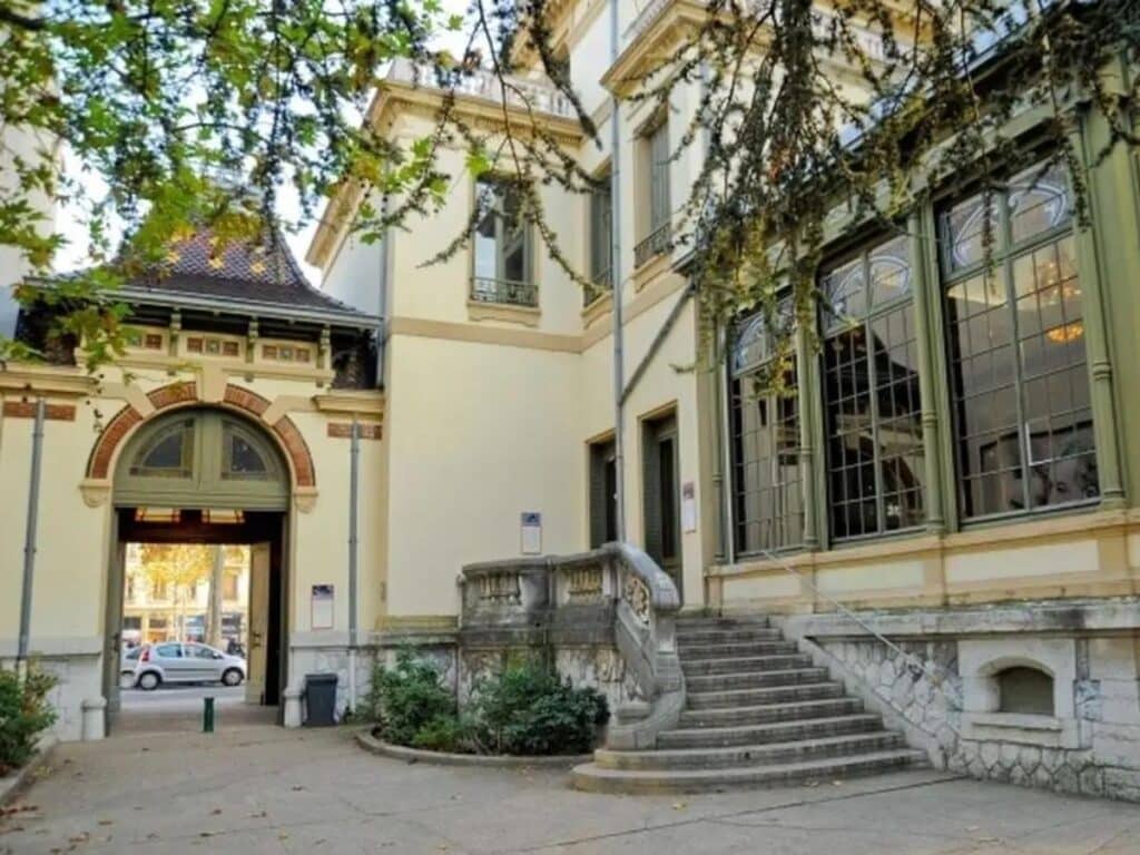 The entrance to the Musée Lumière in Monplaisir, Lyon, features a classic facade with an arched entryway, large windows, and a stone staircase, nestled among green foliage, inviting a glimpse into the cinematic past of the city.