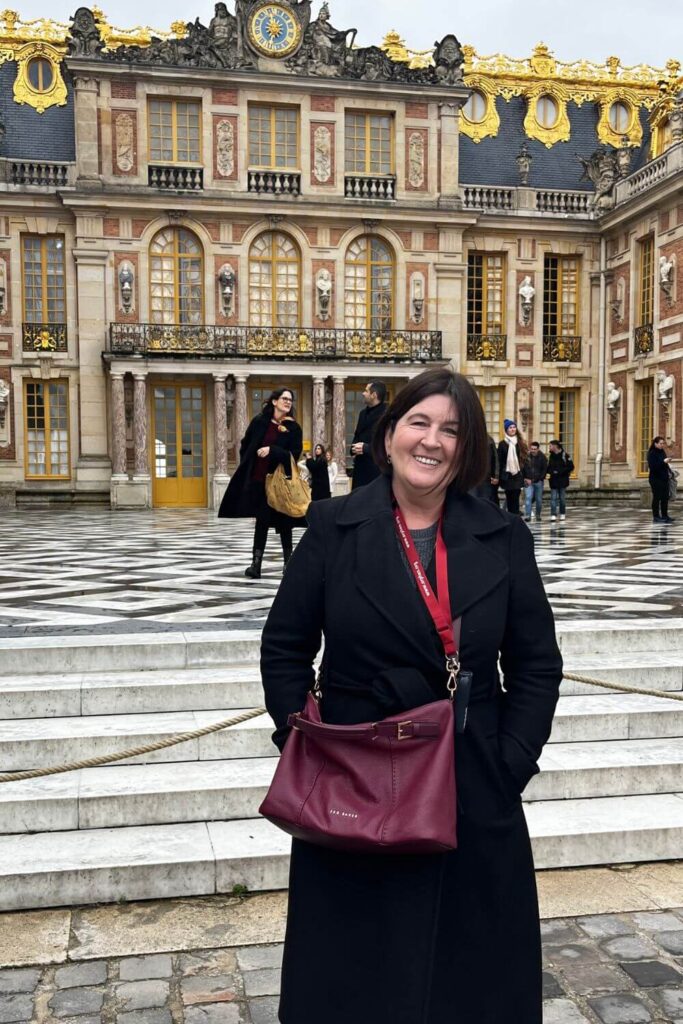 A cheerful woman in business attire, with a burgundy handbag, standing in front of the black and white marble courtyard at the Palace of Versailles, with visitors and the richly decorated palace facade in the background.