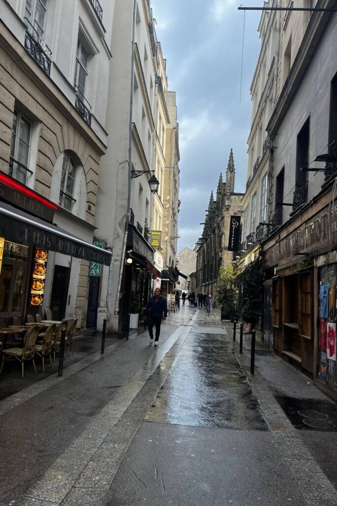 A wet cobblestone street in Paris lined with shops and restaurants, leading towards an ornate Gothic church, capturing the authentic Parisian atmosphere.