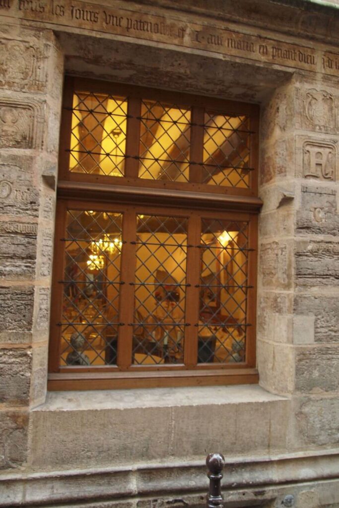 Warm light shines through a leaded glass window set into a historic stone wall with engraved inscriptions, evoking the ambiance of an alchemist's abode in Paris, possibly once related to Nicolas Flamel.