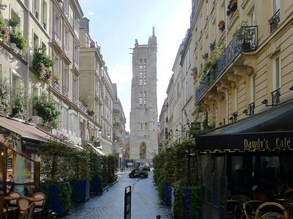 Quaint Parisian street lined with flower-bedecked balconies and outdoor café seating, with a view of the historic Saint-Jacques Tower in the background on a sunny day.