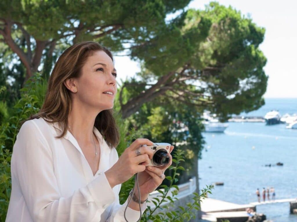 A woman in a white blouse holds a camera and looks upward thoughtfully while standing near lush greenery, with a scenic view of the ocean and yachts in the background.