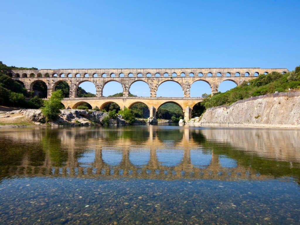 The Pont du Gard, an ancient Roman aqueduct bridge, stands majestically over a tranquil river, its reflection mirroring its grand arches. The clear blue sky and the calm waters accentuate the engineering marvel of this UNESCO World Heritage site.