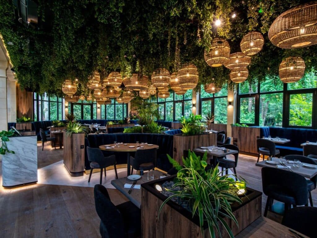 GPT
An elegant restaurant interior, lush with hanging greenery and basket-woven pendant lights, creating a warm and inviting ambiance. The large windows offer a view of green foliage outside, complementing the natural wood furnishings and blue upholstered seating inside.