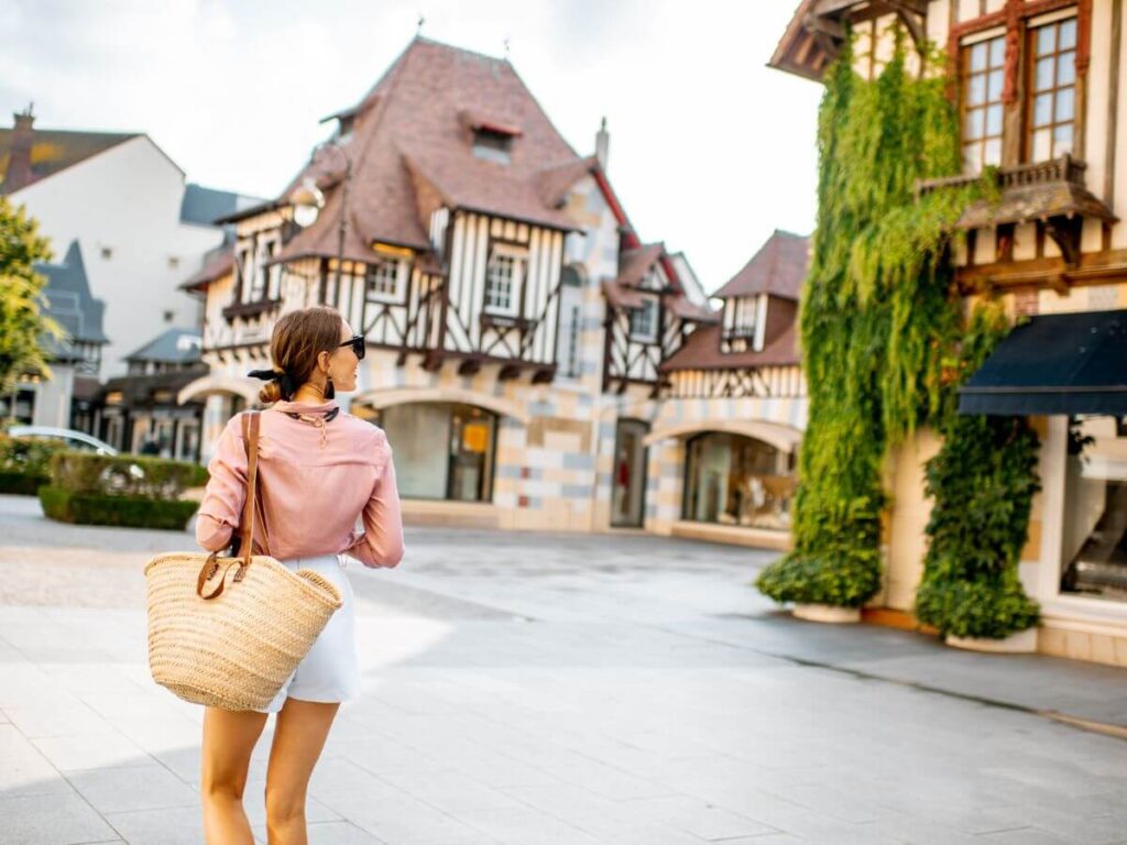 A woman in a casual pink blouse and white shorts carries a woven basket bag while strolling through a quaint French town square, with traditional half-timbered houses adorned with greenery in the background, evoking the charm of rural France