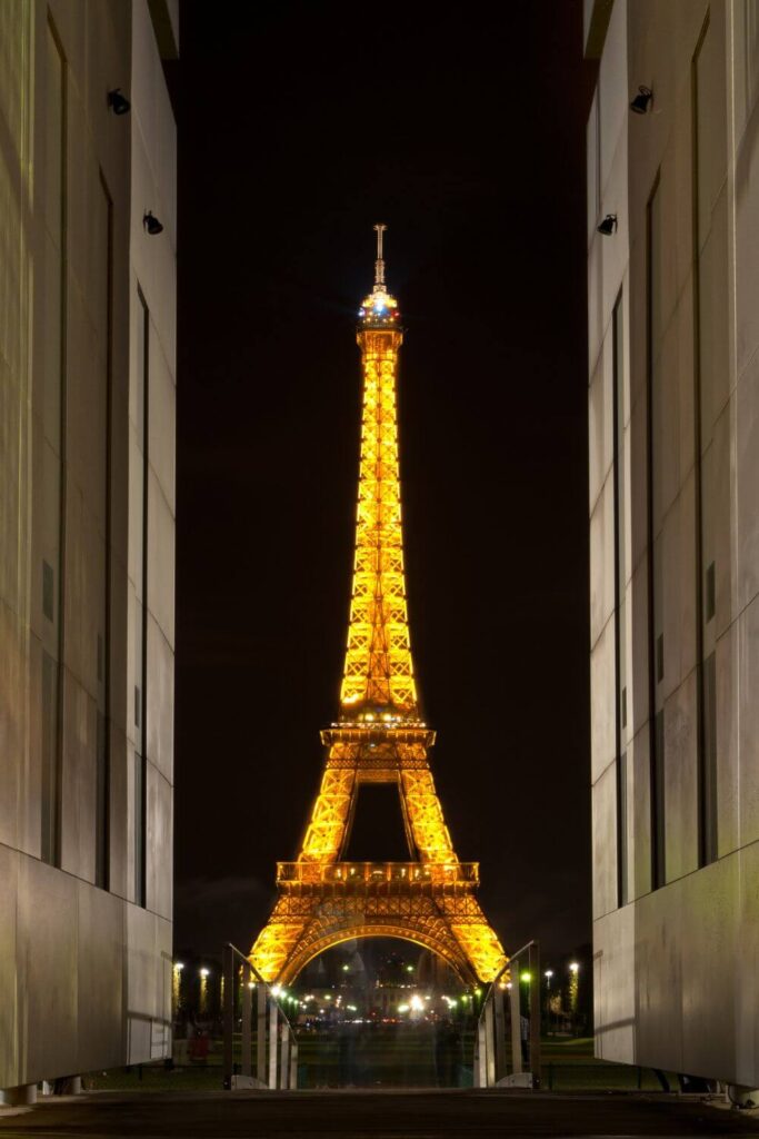 The Eiffel Tower captured at night, glowing with golden light, framed symmetrically by two modern buildings on either side. The pathway leading to the tower is lined with lights, enhancing the geometric beauty of the scene.