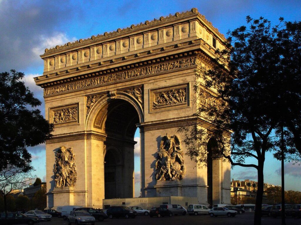 The Arc de Triomphe in Paris, France, captured in the warm glow of the setting sun. The monument's intricate sculptures and friezes are highlighted by the golden sunlight, contrasting with the shadowed foreground. The arch stands majestic against a clear sky, while vehicles are parked and trees frame the scene, adding life and context to this emblem of French history and triumph.