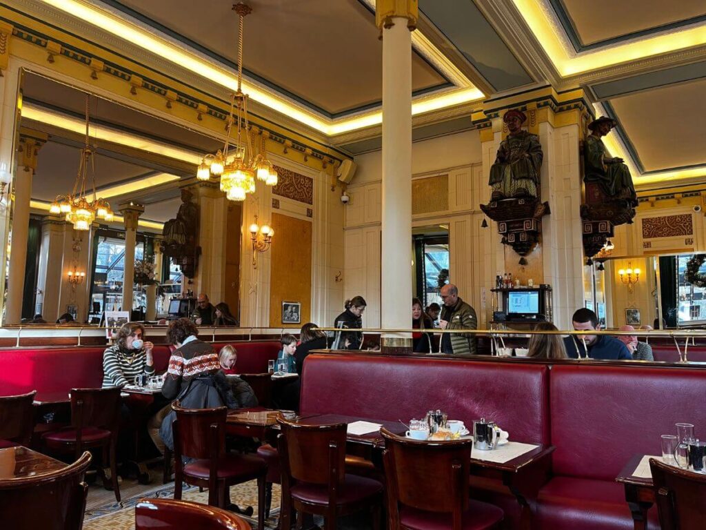 Inside the historically rich Les Deux Magots café, guests enjoy their time surrounded by opulent chandeliers, mirrored columns, and distinctive wooden statues, all under the soft glow of natural light.