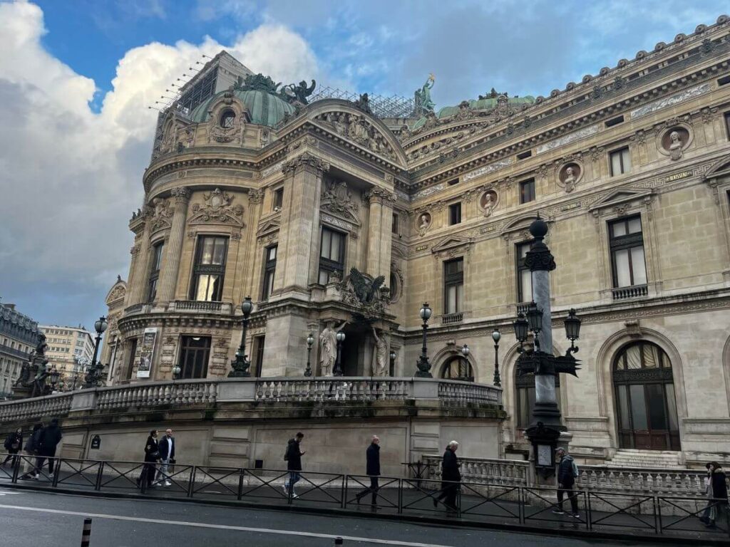 The Palais Garnier, an opera house in Paris, showcasing its elaborate Beaux-Arts architecture with sculptures and ornate detailing. Pedestrians are seen walking by the historic building under a partly cloudy sky.