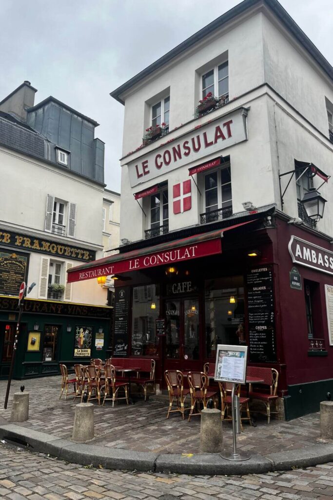 The corner view of 'Le Consulat,' a traditional Parisian cafe with red awnings and outdoor seating on cobblestone streets in Montmartre.