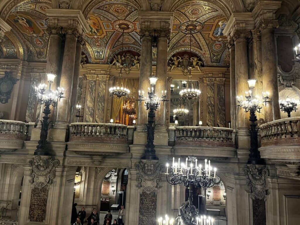 The Grand Foyer of the Palais Garnier with its lavish mosaic flooring, grand marble columns, and ornately decorated ceilings. The space is illuminated by multiple chandeliers, reflecting the opulence of the venue.