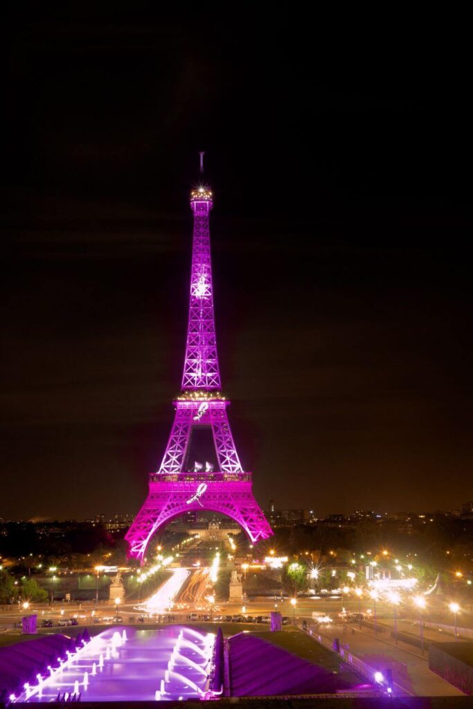 The Eiffel Tower stands tall against the night sky, bathed in vibrant purple lighting. Below, an event with purple illuminated fountains adds to the festive atmosphere. The surrounding park and streets are dotted with the warm glow of streetlights.