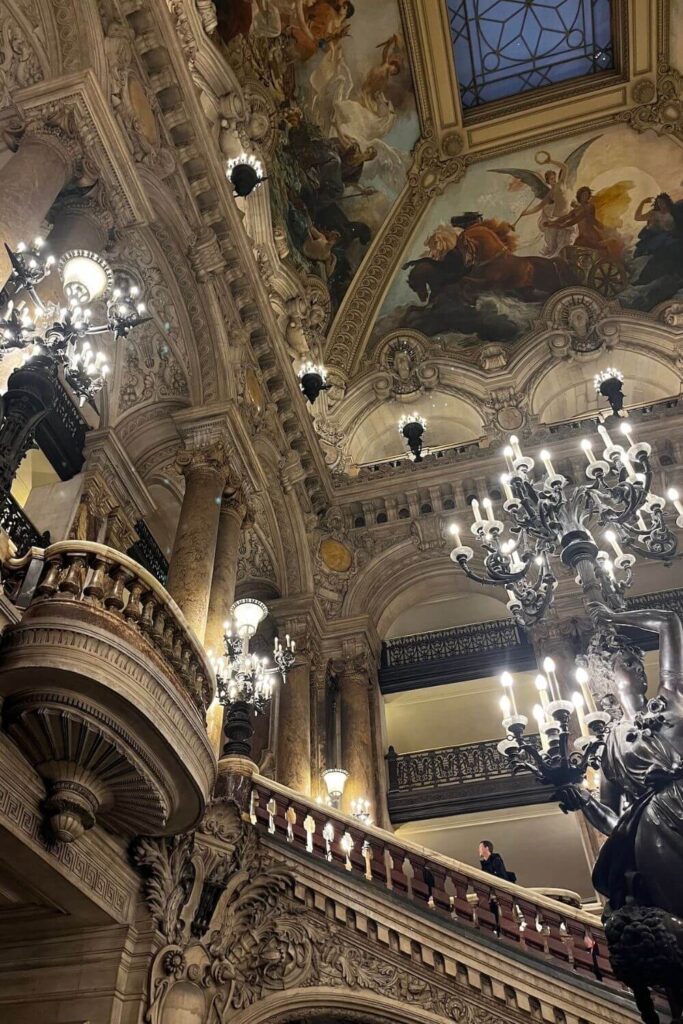 The image highlights the grandeur of the Palais Garnier's interior, featuring the ornate staircase and classical sculptures under the glow of elaborate chandeliers, with a painted ceiling above. The architecture facilitates a luxurious ambience for visitors to admire the surroundings and each other.