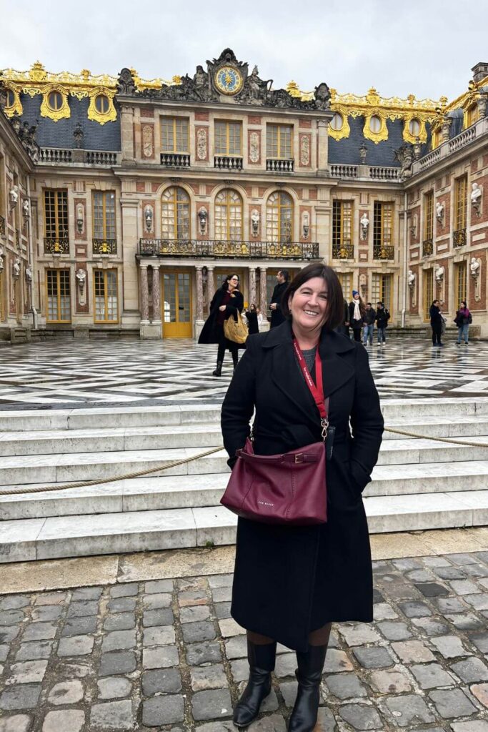 A smiling person standing in front of the ornate golden gates of the Palace of Versailles, with the palace's intricate facade and clock in the background.