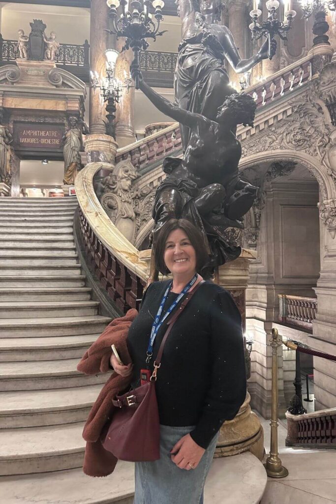 A visitor is pictured smiling on the grand staircase of the Palais Garnier, with a sculptural bronze lamp post and ornate balustrades in the background, capturing the elegance of this historic opera house.