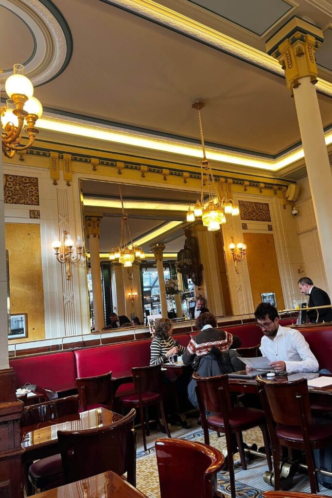 Patrons seated at red banquettes inside the elegant Les Deux Magots café, with golden chandeliers and classical columns reflecting in the mirrored walls, embodying the quintessential Parisian café culture.