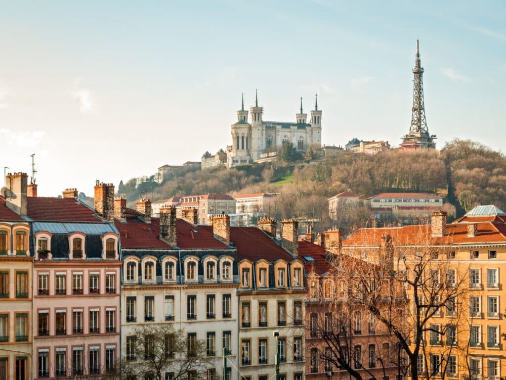 A scenic view of Lyon with a layered composition of colorful facades in the foreground and the iconic Notre-Dame de Fourvière Basilica and a resembling structure of the Eiffel Tower in the distance, set against a soft evening sky.