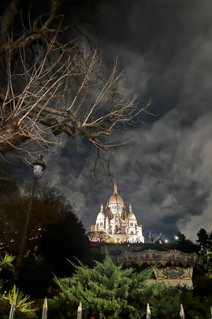 Night view of the illuminated Sacré-Cœur Basilica seen through bare tree branches with a dramatic cloudy sky and a street lamp in the foreground.