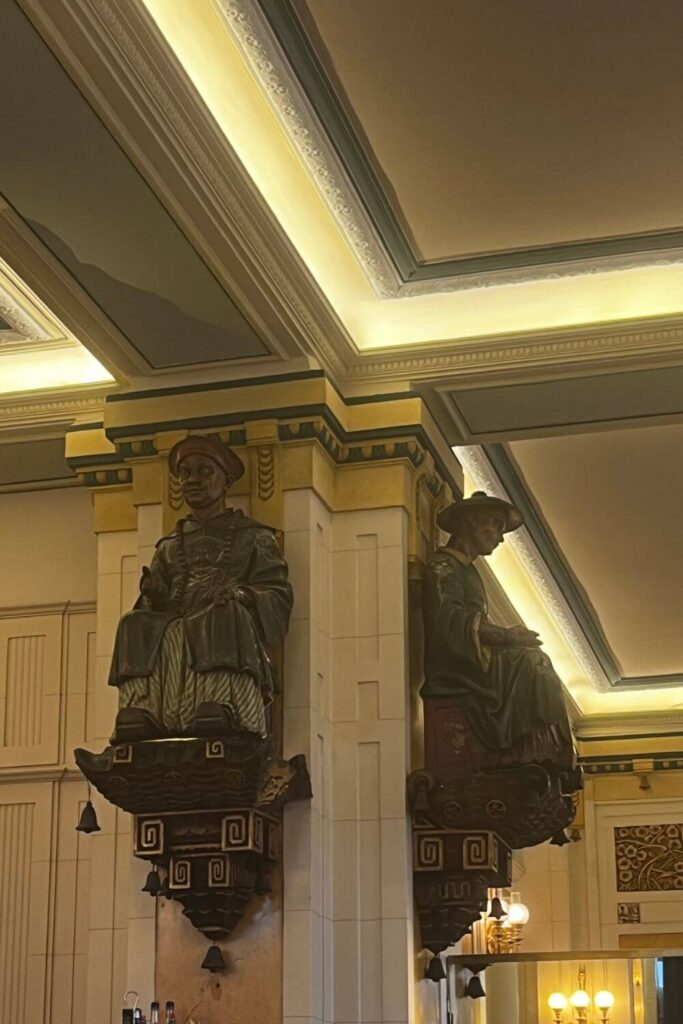 Interior view of Les Deux Magots café, showcasing ornate wooden figurines perched on the wall, with elegant lighting that accentuates the detailed ceiling and classical architectural elements.