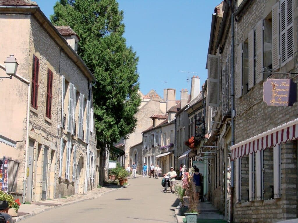 GPT
A tranquil street in Vézelay lined with traditional stone buildings, each adorned with shutters and signs offering local wares, creates a picturesque scene of rural French life under a clear blue sky. The street is dotted with leisurely strolling visitors, adding to the charm of this historic village.